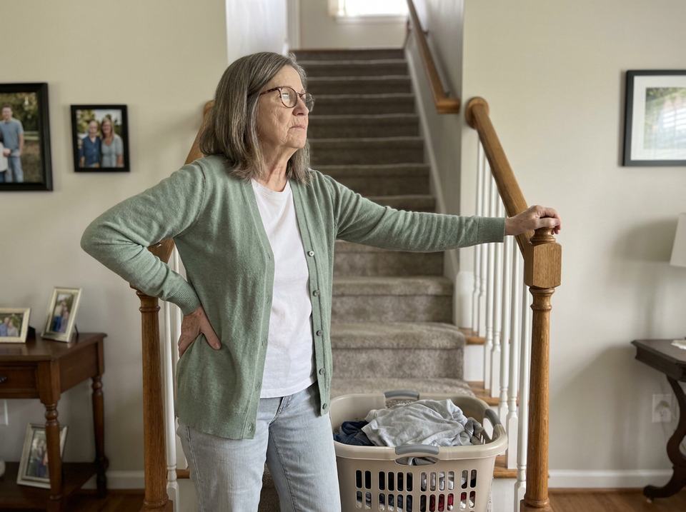 Woman in her 60s at the bottom of her staircase with a laundry basket, holding the side of her hip