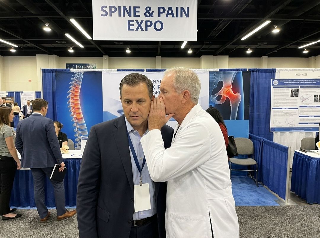 A surgeon whispering to a sales rep at a spine and pain expo